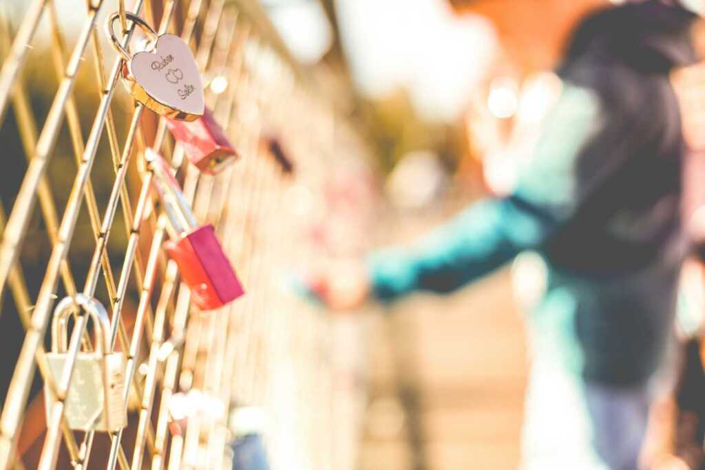 A fence with many locks on it and a blurred person in the background attaching another