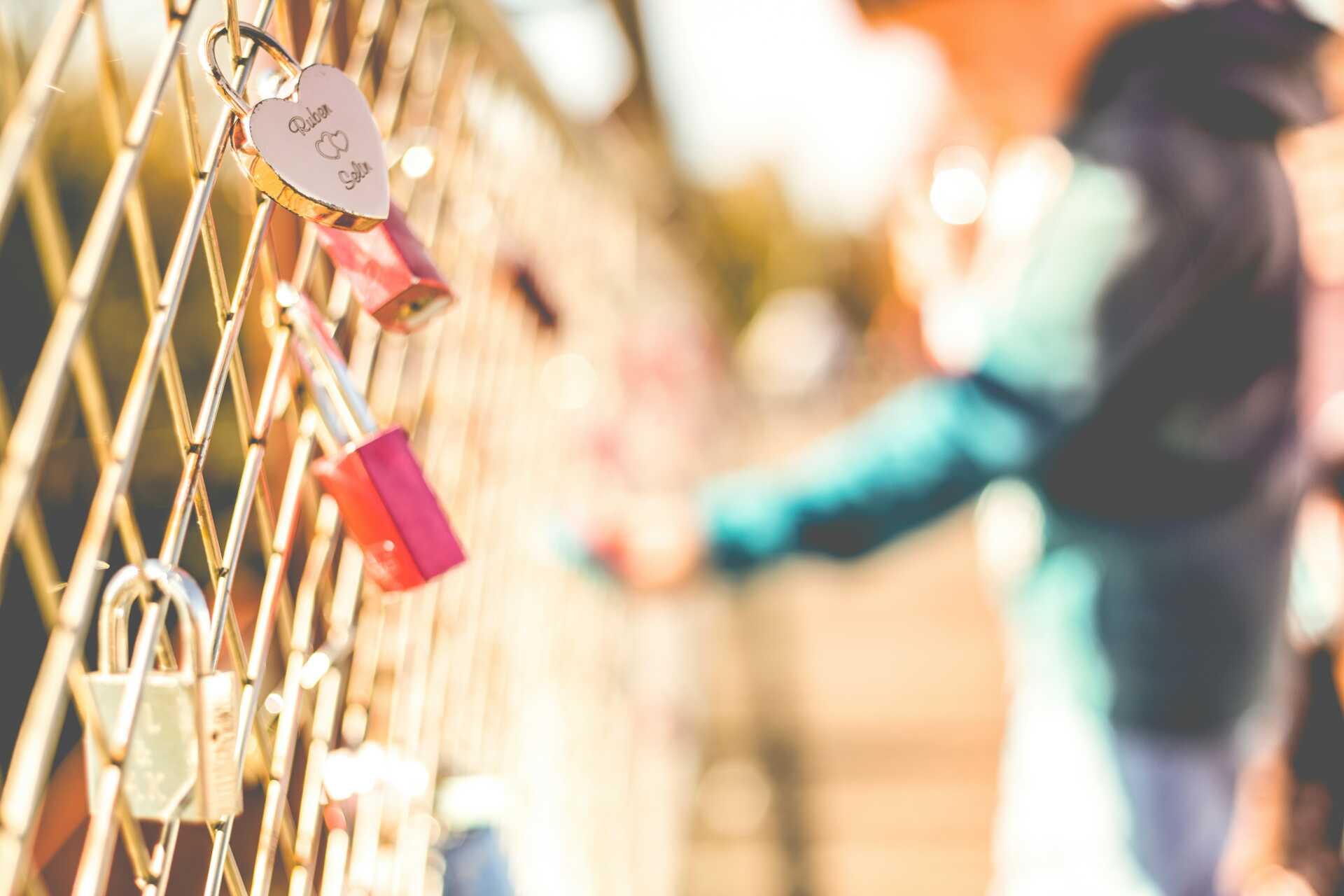 A fence with many locks on it and a blurred person in the background attaching another