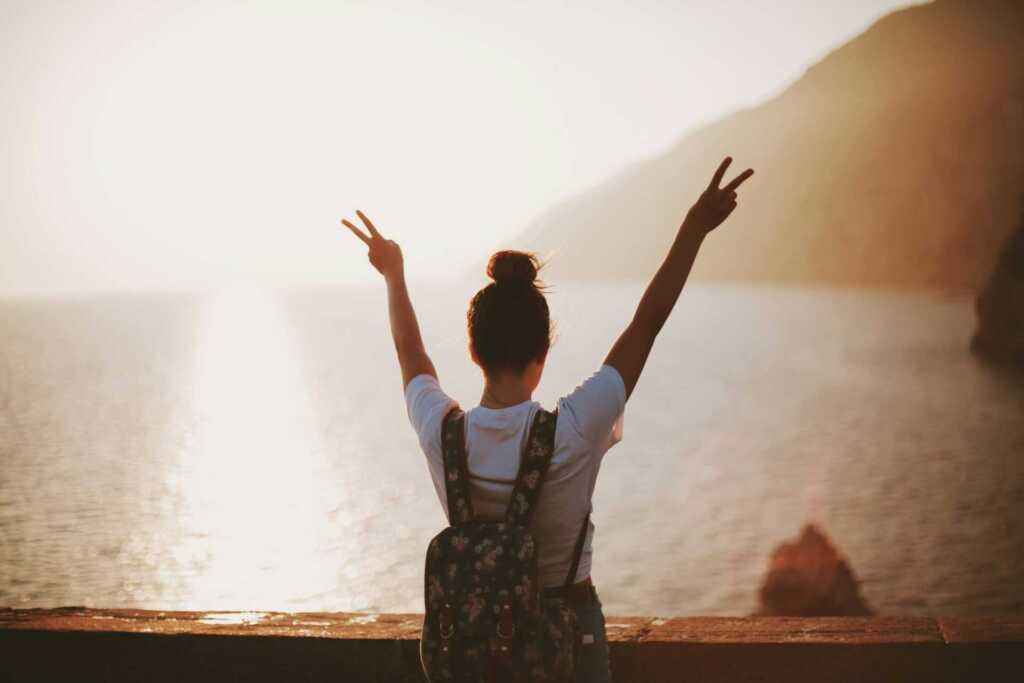 A women looking out over a lake with the sun shining, holding up her hands in two peace signs