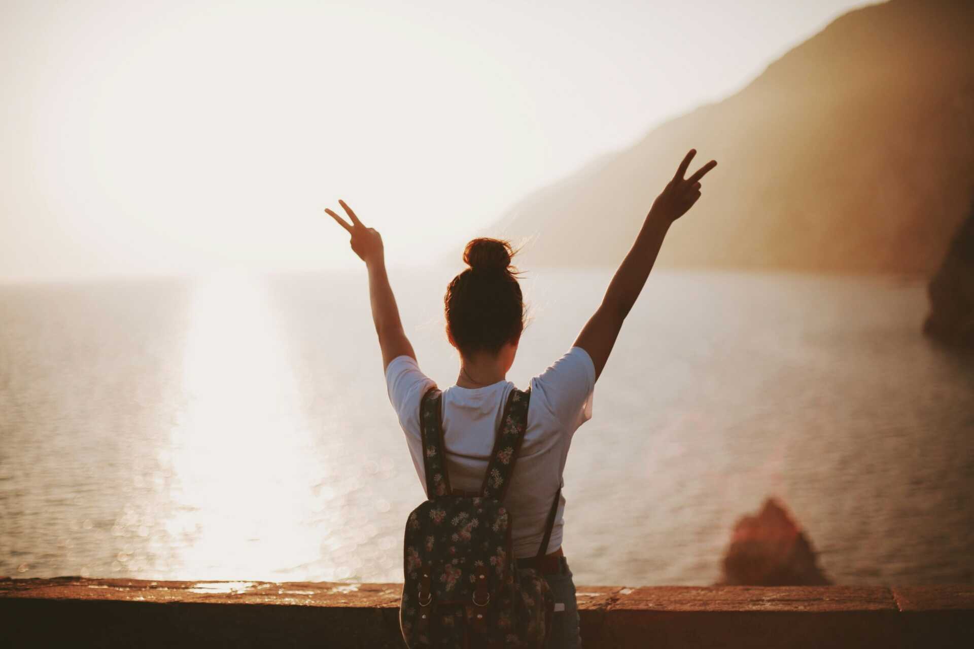 nine koepfer d2 H9 WSl7o unsplash A women looking out over a lake with the sun shining, holding up her hands in two peace signs