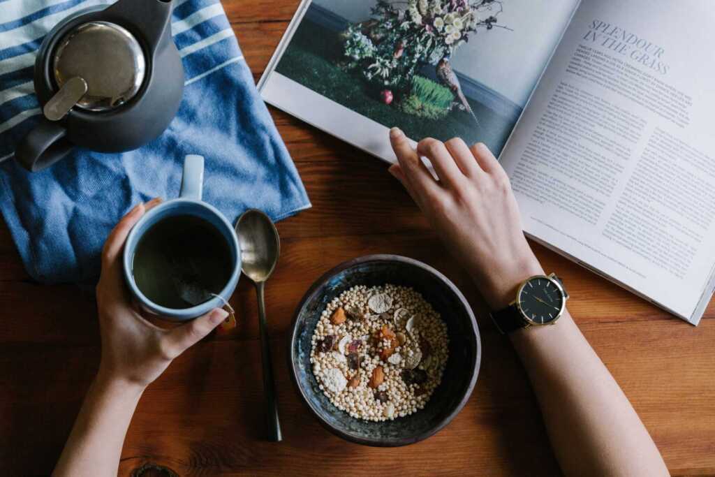 overhead view of a kitchen table where someone is reading a book, drinking coffee, and has a bowl of oatmeal in front of them