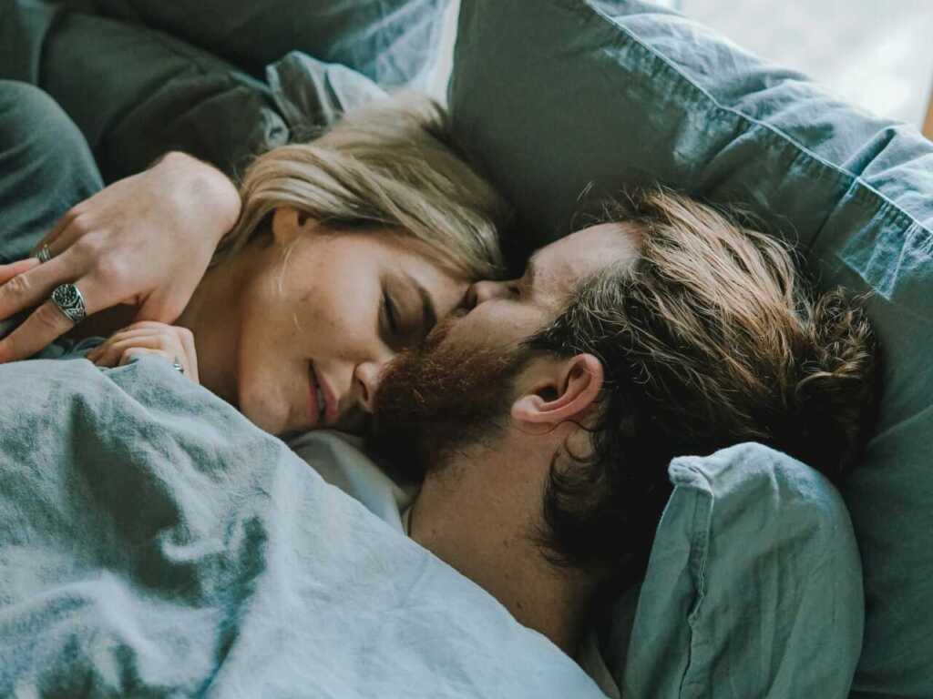 A couple cuddling in bed under the covers, with the male partner kissing the female partner on the forehead