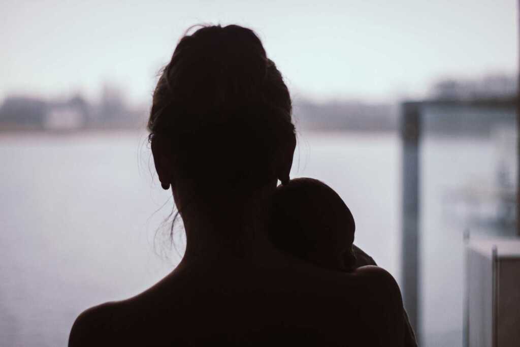 A black and white photo of a woman holding her baby and looking out onto the water