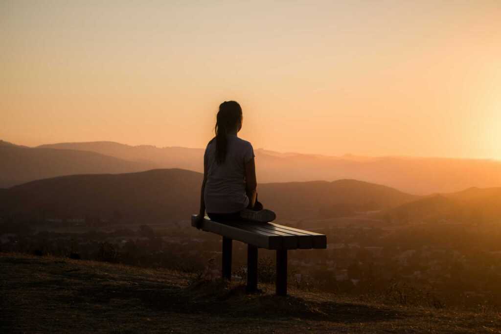 A woman sitting on a bench looking out over the hills at sunset