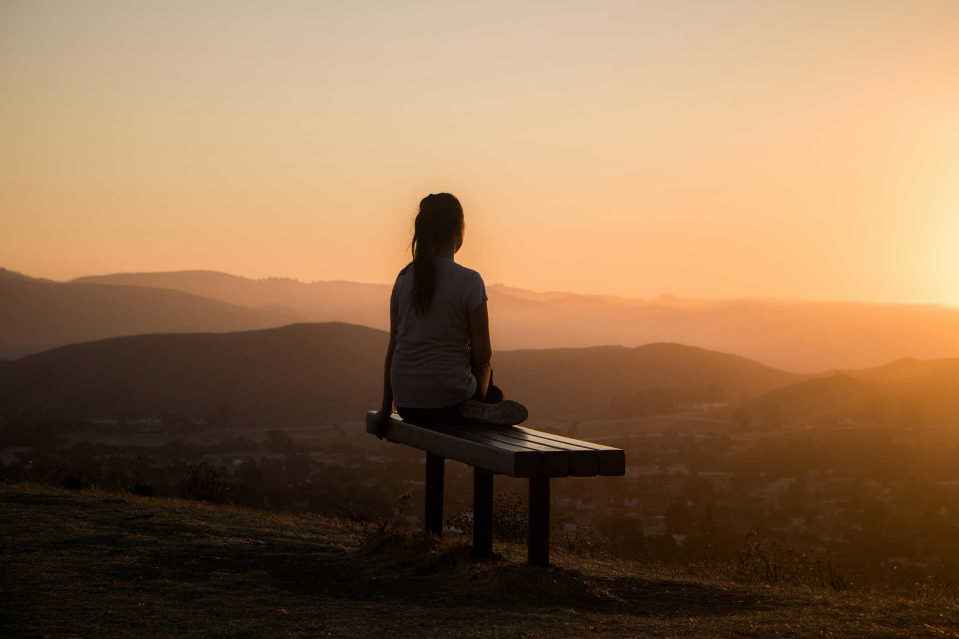 A woman sitting on a bench looking out over the hills at sunset